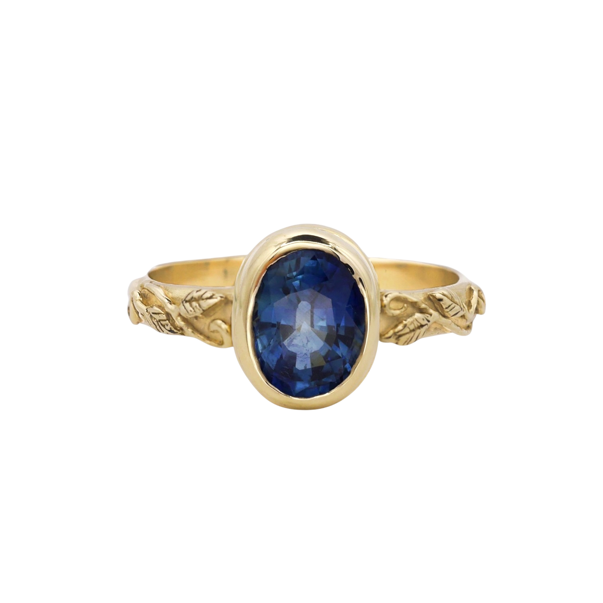 Interlaced Ring Sapphire — A deep-blue oval sapphire is bezel-set above a yellow-gold band carved with leaves and curling vines.