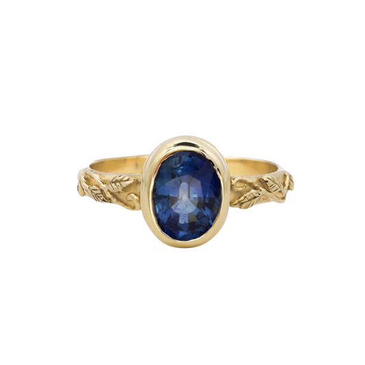 Interlaced Ring Sapphire — A deep-blue oval sapphire is bezel-set above a yellow-gold band carved with leaves and curling vines.