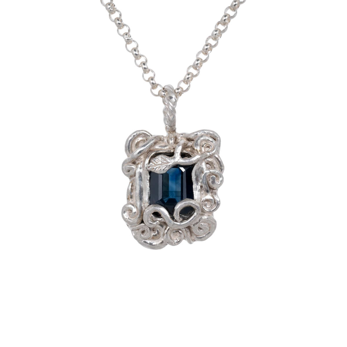 Sapphire Amulet Necklace Silver — A deep-blue rectangular sapphire sits within an ornate silver-colored amulet of curling vines and leaf relief.