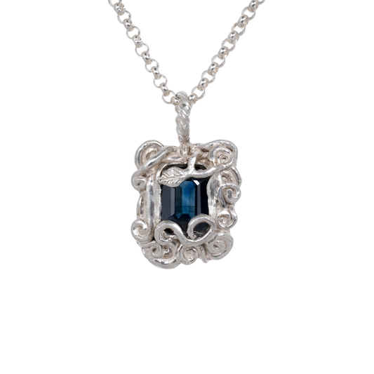 Sapphire Amulet Necklace Silver — A deep-blue rectangular sapphire sits within an ornate silver-colored amulet of curling vines and leaf relief.