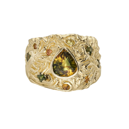 The Turning Art Ring — A wide yellow-gold-colored statement ring centers an olive-and-gold pear-shaped stone in a bezel amid nine visible multicolor accent stones and dense raised scrollwork.