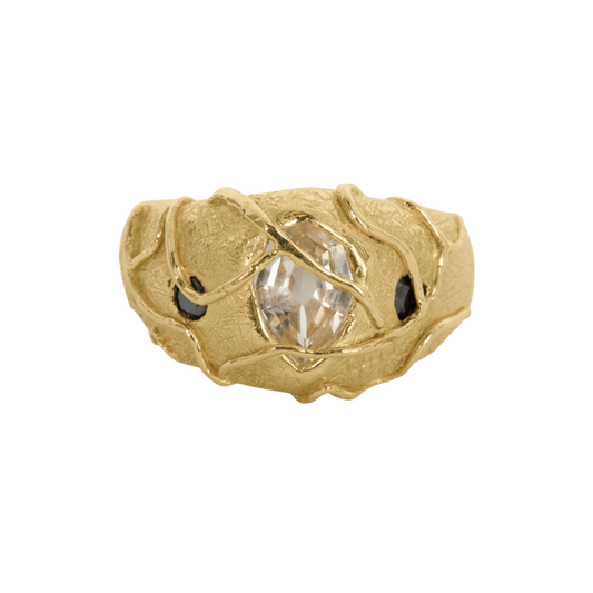 Watch Your Step Ring — A wide textured yellow-gold-colored ring holds one elongated colorless center stone and two small black pear-shaped side stones beneath crossing vine-like prongs.